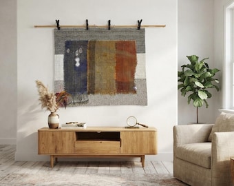 Puede incluir: Un tapiz rectangular para pared con bloques de color verticales en azul marino, amarillo mostaza y naranja quemado, sobre un fondo gris y blanco. El tejido está suspendido de una barra de madera mediante cuatro clips de metal negro.