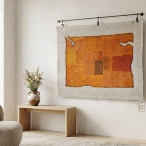 Op de afbeelding: Een rechthoekige wandhanger met een patchwork van oranje, roestkleurige en bruine stoffen vierkanten genaaid op een lichtbeige linnen achtergrond. Het textiel hangt aan een donkere metalen roede met drie clips en heeft zichtbare donkere stiksels.