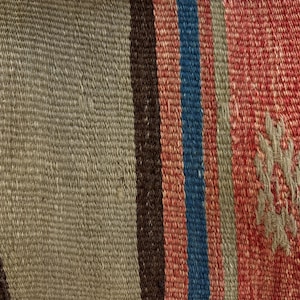 Op de afbeelding: Een geweven textiel met verticale strepen in tinten olijfgroen, donkerbruin, blauw en rood. De stof heeft een gestructureerde platgeweven constructie met een klein, lichtgekleurd geometrisch motief geborduurd op het rode gedeelte.