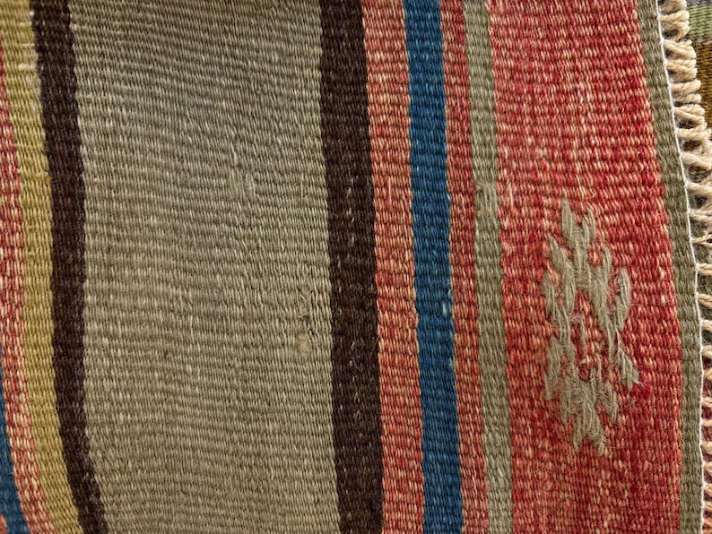 Op de afbeelding: Een geweven textiel met verticale strepen in tinten olijfgroen, donkerbruin, blauw en rood. De stof heeft een gestructureerde platgeweven constructie met een klein, lichtgekleurd geometrisch motief geborduurd op het rode gedeelte.