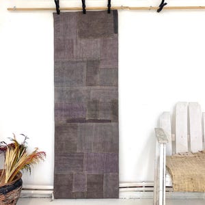 Peut inclure: Une tenture murale rectangulaire présentant un motif en patchwork composé de divers rectangles de tissu dans des tons gris et violet sourd. Le textile est suspendu à une tringle en bois par des pinces noires, offrant un aspect texturé.