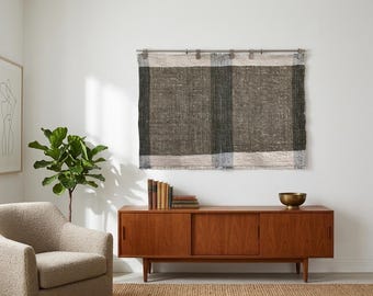 May include: A rectangular wall hanging featuring a woven textile with a large-scale plaid pattern in shades of dark charcoal, grey, and off-white. The fabric has a visible coarse texture and frayed edges, suspended from a metal rod with clips.