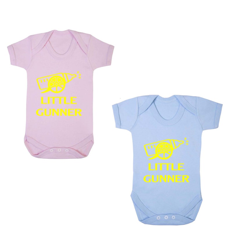 May include: Two baby onesies, one pink and one light blue, with a yellow graphic of a cannon and the text "Little Gunner".