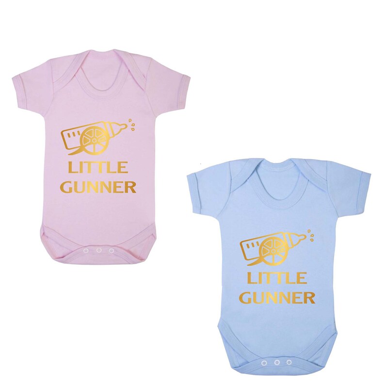 May include: Two baby onesies, one pink and one blue, with a gold graphic of a baby bottle with a cannon on it. The text "Little Gunner" is printed in gold below the graphic.