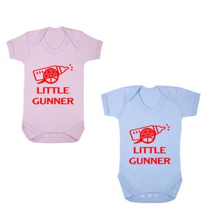 May include: Two baby onesies, one pink and one light blue, with a red graphic of a baby bottle shaped like a cannon with the text "Little Gunner" on each.