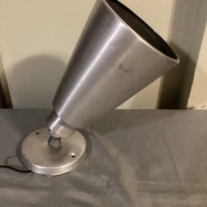 May include: A brushed metal spotlight with a conical shade and a circular base. The lamp is angled upwards, showcasing its design. The base has three screw holes. The lamp is silver in color.