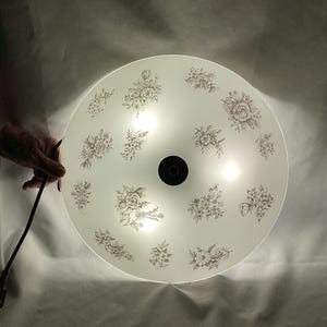 May include: A white ceiling light fixture with a floral design. The light fixture has a round, frosted glass shade with a black metal center.