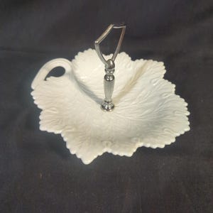 May include: White leaf-shaped serving dish with a silver handle. The dish has a detailed embossed leaf pattern and a curved handle for easy carrying. The dish is made of ceramic or porcelain and is suitable for serving appetizers or desserts.