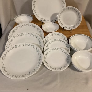May include: A collection of white Corelle dinnerware with a gray floral vine pattern around the rims. The set includes various sized plates and bowls, arranged on a wooden surface.