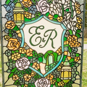 May include: Stained glass panel featuring a central shield with the letters "E&R" in green script. The panel is framed by colorful floral and leaf designs in shades of yellow, green, and white, with two yellow lanterns.