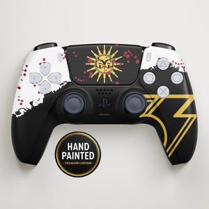 Op de afbeelding: Handgeschilderde PlayStation 5-controller in zwart en wit, met gouden en rode details. Het ontwerp bevat een zon- en bliksemmotief. De controller heeft een sticker met de tekst "HAND PAINTED - PREMIUM CUSTOM".