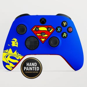 Op de afbeelding: Een blauwe Xbox-controller met een Superman-logo en een geel en zwarte Superman-afbeelding aan de linkerkant. De controller heeft zwarte knoppen en een rode onderkant. Een ronde sticker leest "HAND PAINTED - PREMIUM CUSTOM."
