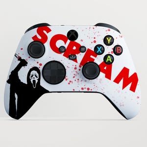 Op de afbeelding: Een witte draadloze videogamecontroller met een zwart silhouet van het Ghostface-personage dat een mes vasthoudt aan de linkerkant, het woord SCREAM in grote rode letters in het midden en rode bloedspatten over de gehele behuizing.