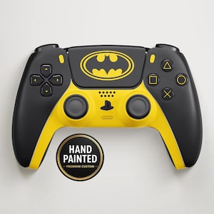 Puede incluir: Un mando de juego inalámbrico negro y amarillo con un destacado logotipo de Batman en amarillo sobre el panel táctil central. El mando incluye un emblema circular con el texto HAND PAINTED PREMIUM CUSTOM. Los botones y joysticks son negros.