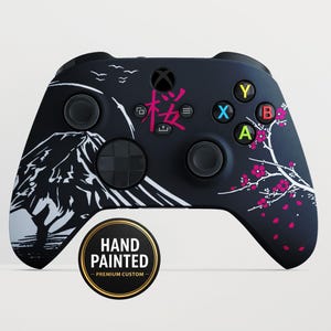 Op de afbeelding: Zwarte Xbox-controller met een aangepast ontwerp met een berglandschap en kersenbloesems. De controller heeft roze Japanse tekst en de woorden "HAND PAINTED - PREMIUM CUSTOM" op een goudzwarte sticker.
