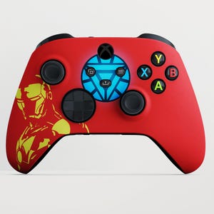 Może przedstawiać: Czerwony kontroler Xbox z żółto-czerwonym motywem Iron Mana po lewej stronie. Kontroler ma czarne przyciski i niebieski podświetlany centralny wzór. Przyciski funkcyjne są zielone, żółte, niebieskie i czerwone.