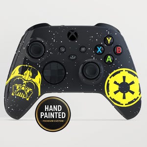 Puede incluir: Mando de Xbox negro con un diseño pintado a mano que presenta a Darth Vader en amarillo y un símbolo imperial amarillo. El mando tiene motas blancas y el texto "HAND PAINTED - PREMIUM CUSTOM" en una pegatina circular.