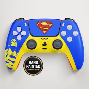 Könnte beinhalten: Ein blau, gelb und rot handbemalter PlayStation 5-Controller mit dem Superman-Logo. Der Controller hat eine gelbe Mitte, blaue Seiten und rote Akzente. Das Superman-Logo ist prominent in der Mitte oben abgebildet. Ein runder Aufkleber lautet "Handbemalt - Premium Custom."