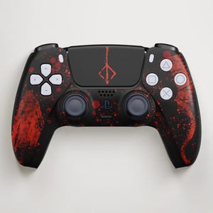 May include: Black PlayStation 5 controller with a red splatter design. The controller features white buttons and a red emblem on the touchpad. The design covers the handles and the face of the controller.