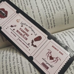 Może przedstawiać: R&oacute;żowy i czarny bilet z tekstem "Dożywotnie członkostwo w Vampire Bookclub" i czerwonym sercem z nietoperzowymi skrzydłami. Bilet ma kod kreskowy i tekst "wino czerwone w cenie".