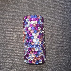 May include: A cylindrical object covered in colorful rhinestones. The rhinestones are in shades of blue, purple, pink, and clear, creating a sparkling effect. The object is likely a decorative item.