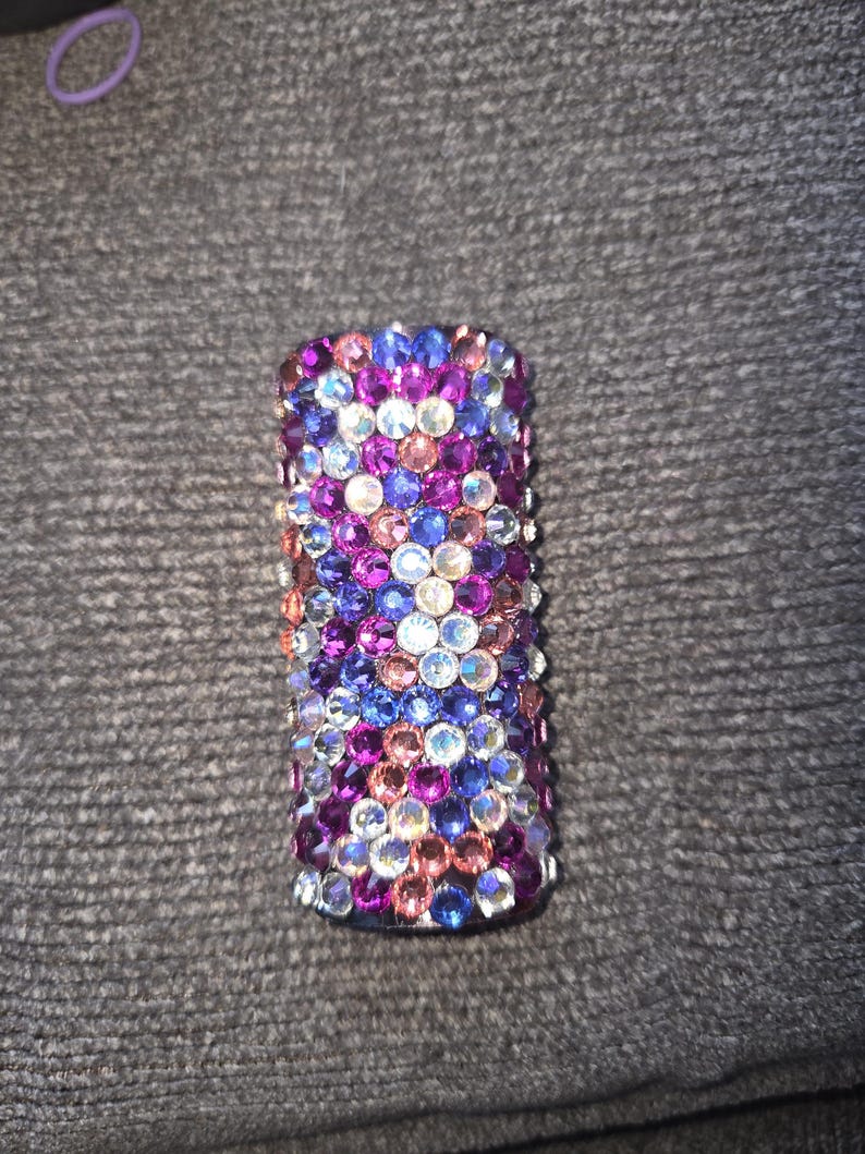 May include: A cylindrical object covered in colorful rhinestones. The rhinestones are in shades of blue, purple, pink, and clear, creating a sparkling effect. The object is likely a decorative item.