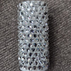 May include: A cylindrical, white object fully encrusted with numerous, sparkling, clear rhinestones. The rhinestones are closely set, creating a dazzling, textured surface. The object has a solid, opaque base.