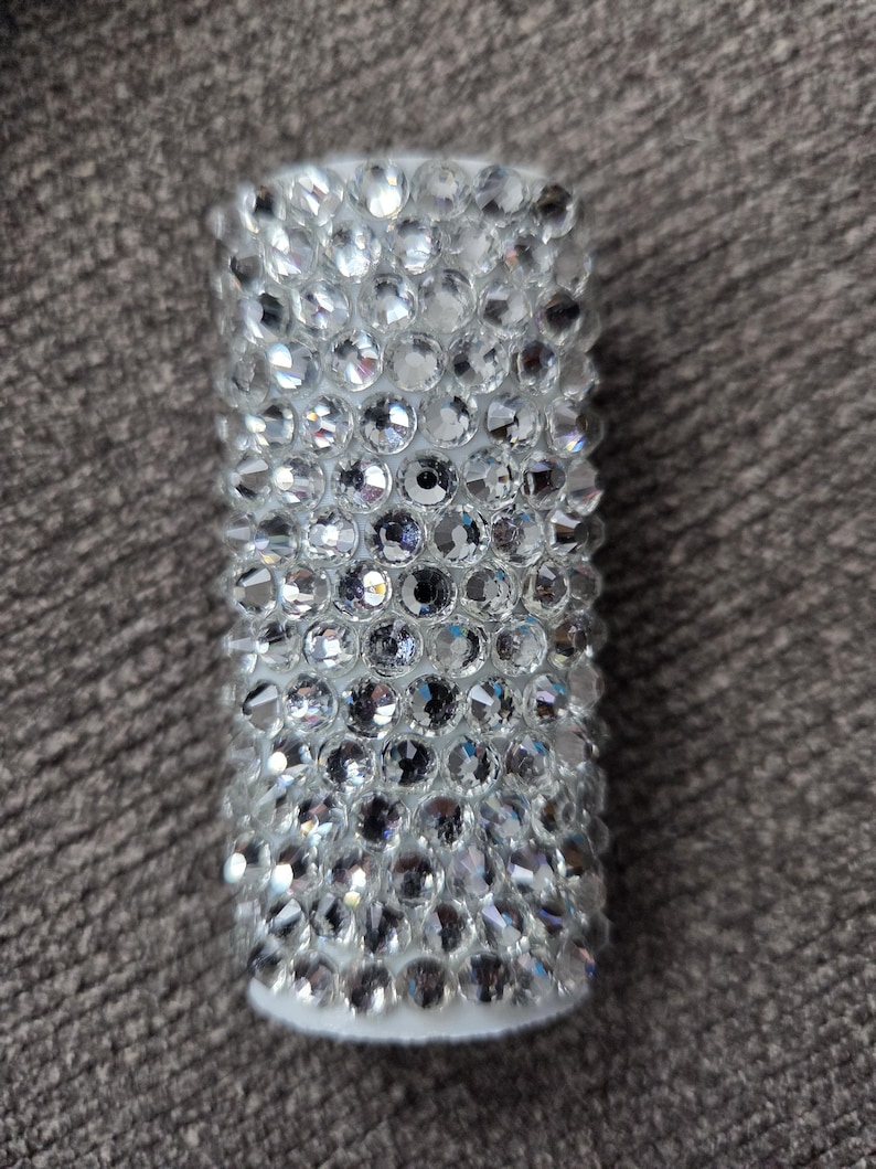 May include: A cylindrical, white object fully encrusted with numerous, sparkling, clear rhinestones. The rhinestones are closely set, creating a dazzling, textured surface. The object has a solid, opaque base.