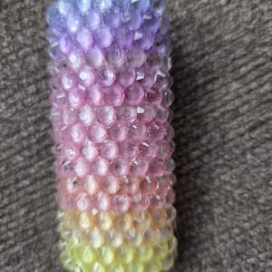 May include: A cylindrical candle with a gradient of colors. The top is purple, transitioning to pink, then yellow at the base. The surface is textured with small, round, faceted details.