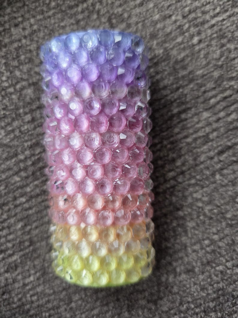 May include: A cylindrical candle with a gradient of colors. The top is purple, transitioning to pink, then yellow at the base. The surface is textured with small, round, faceted details.
