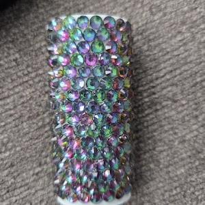 May include: A cylindrical object covered in iridescent, multi-colored rhinestones. The rhinestones are primarily green, purple, and blue, and are arranged closely together. The base of the object is white.