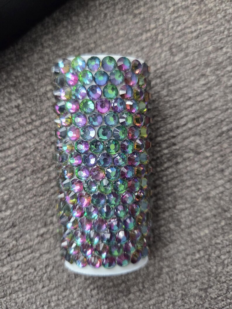 May include: A cylindrical object covered in iridescent, multi-colored rhinestones. The rhinestones are primarily green, purple, and blue, and are arranged closely together. The base of the object is white.