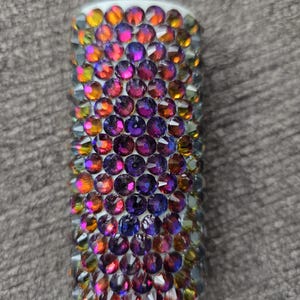 May include: A cylindrical object covered in iridescent, colorful rhinestones. The rhinestones are primarily purple, orange, and yellow, and are arranged closely together. The object has a white base and is set against a gray background.