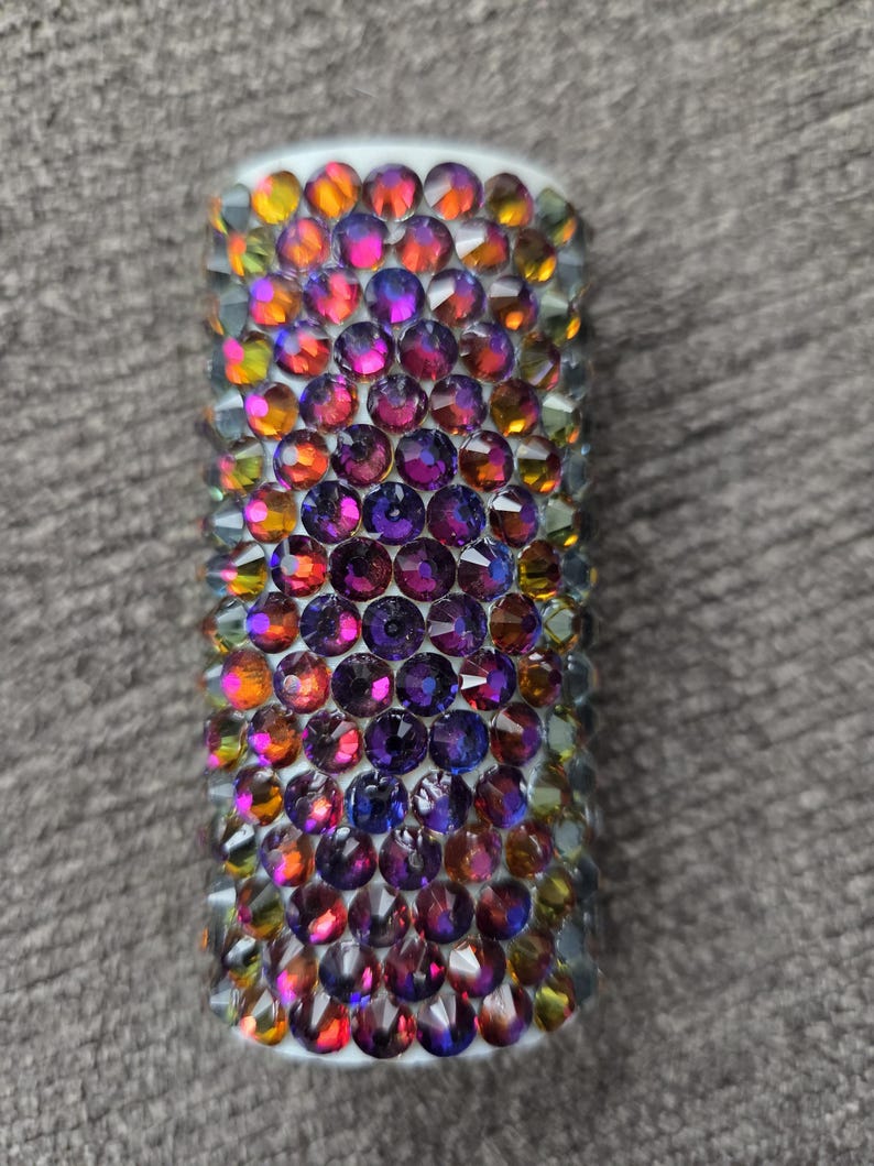 May include: A cylindrical object covered in iridescent, colorful rhinestones. The rhinestones are primarily purple, orange, and yellow, and are arranged closely together. The object has a white base and is set against a gray background.