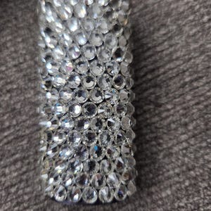 May include: A rectangular, cylindrical object fully encrusted with clear, sparkling rhinestones. The rhinestones are closely set, creating a dazzling, reflective surface. The object appears to be a decorative accessory or a small container.
