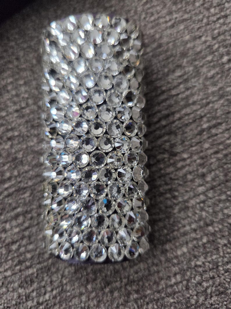 May include: A rectangular, cylindrical object fully encrusted with clear, sparkling rhinestones. The rhinestones are closely set, creating a dazzling, reflective surface. The object appears to be a decorative accessory or a small container.