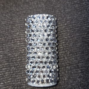 May include: Cylindrical object covered in numerous clear, sparkling rhinestones. The rhinestones are closely set, creating a shimmering, textured surface. The base appears to be white. The object is likely a decorative item or accessory.