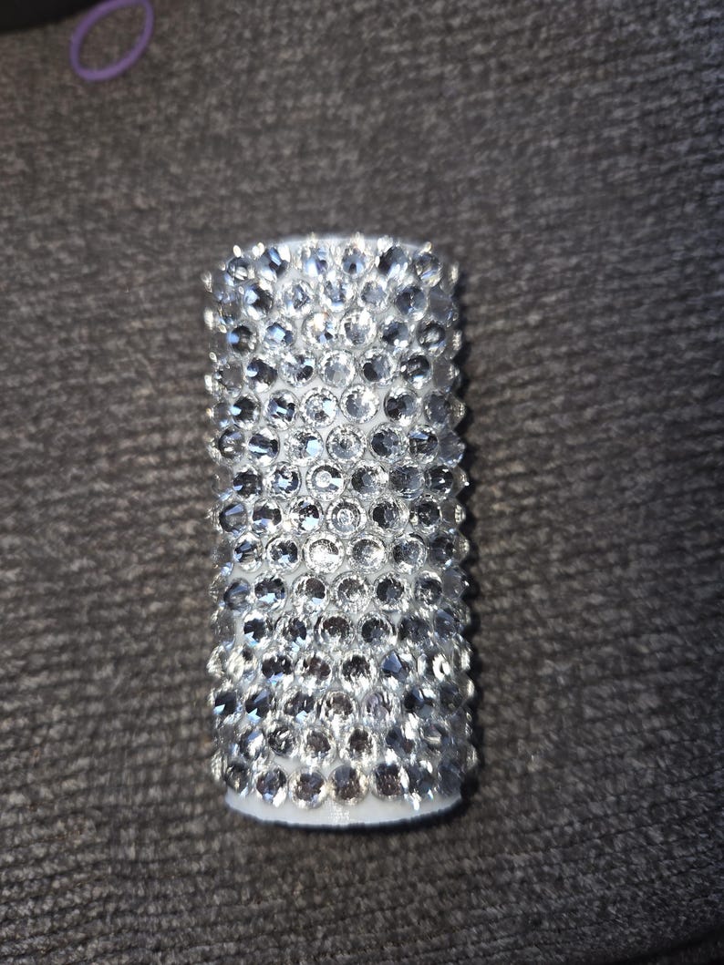May include: Cylindrical object covered in numerous clear, sparkling rhinestones. The rhinestones are closely set, creating a shimmering, textured surface. The base appears to be white. The object is likely a decorative item or accessory.