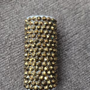 May include: A cylindrical object covered in gold-colored, faceted rhinestones. The top and bottom edges are white. The rhinestones are closely packed together, creating a textured surface. The object is likely a decorative item.