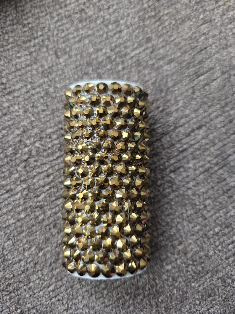 May include: A cylindrical object covered in gold-colored, faceted rhinestones. The top and bottom edges are white. The rhinestones are closely packed together, creating a textured surface. The object is likely a decorative item.