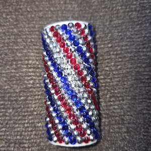May include: A cylindrical object covered in red, silver, and blue rhinestones. The rhinestones are arranged in a pattern of diagonal stripes. The object has a white base and is likely a decorative item.