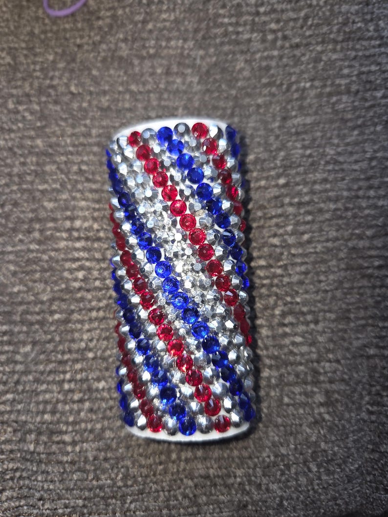 May include: A cylindrical object covered in red, silver, and blue rhinestones. The rhinestones are arranged in a pattern of diagonal stripes. The object has a white base and is likely a decorative item.