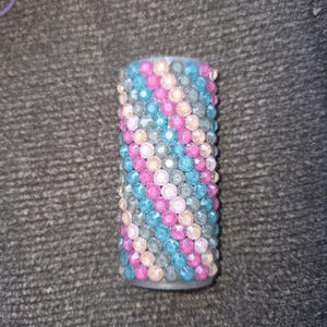 May include: A cylindrical object covered in rows of colorful, sparkling rhinestones. The rhinestones are arranged in diagonal rows of pink, blue, and gray. The object is likely a decorative item or accessory.