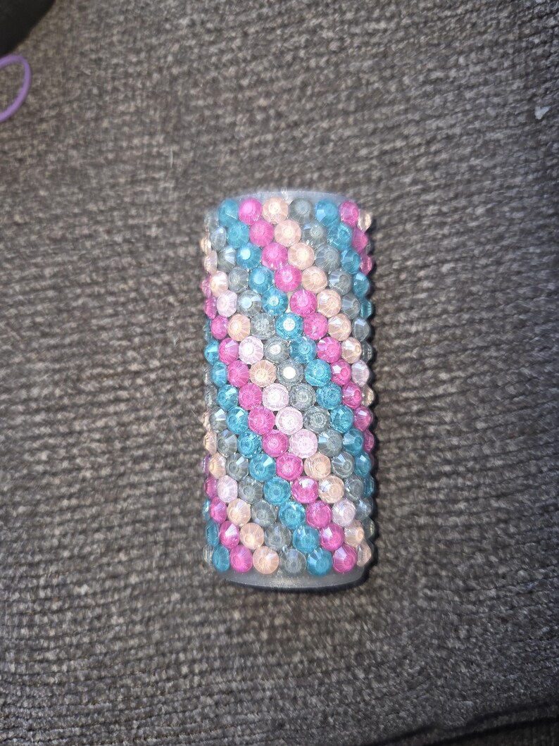 May include: A cylindrical object covered in rows of colorful, sparkling rhinestones. The rhinestones are arranged in diagonal rows of pink, blue, and gray. The object is likely a decorative item or accessory.