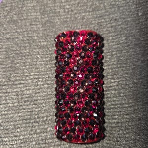 May include: A cylindrical object covered in small, sparkling red and dark purple crystals. The crystals are densely packed, creating a textured surface. The object is set against a gray background.