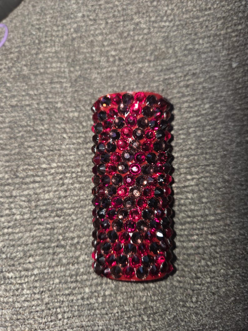 May include: A cylindrical object covered in small, sparkling red and dark purple crystals. The crystals are densely packed, creating a textured surface. The object is set against a gray background.