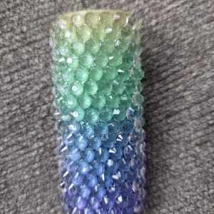 May include: A cylindrical candle with a gradient of colors, transitioning from yellow-green at the top to blue and purple at the bottom. The candle is covered in small, faceted beads.