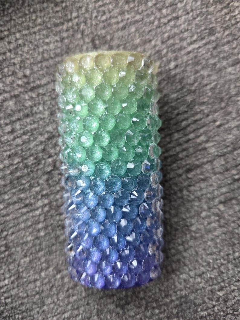 May include: A cylindrical candle with a gradient of colors, transitioning from yellow-green at the top to blue and purple at the bottom. The candle is covered in small, faceted beads.