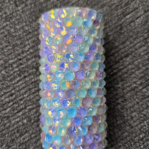 May include: A cylindrical object covered in iridescent, multi-colored rhinestones. The rhinestones are primarily blue, purple, and yellow, and are arranged closely together. The base of the object is white.