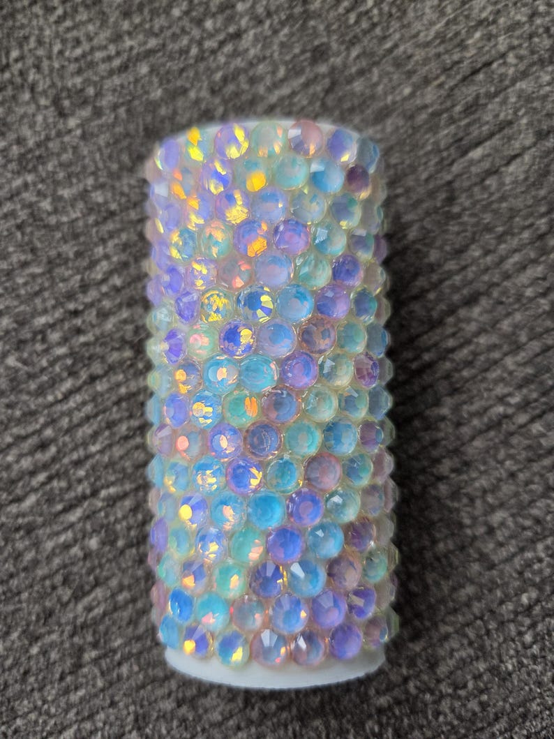 May include: A cylindrical object covered in iridescent, multi-colored rhinestones. The rhinestones are primarily blue, purple, and yellow, and are arranged closely together. The base of the object is white.